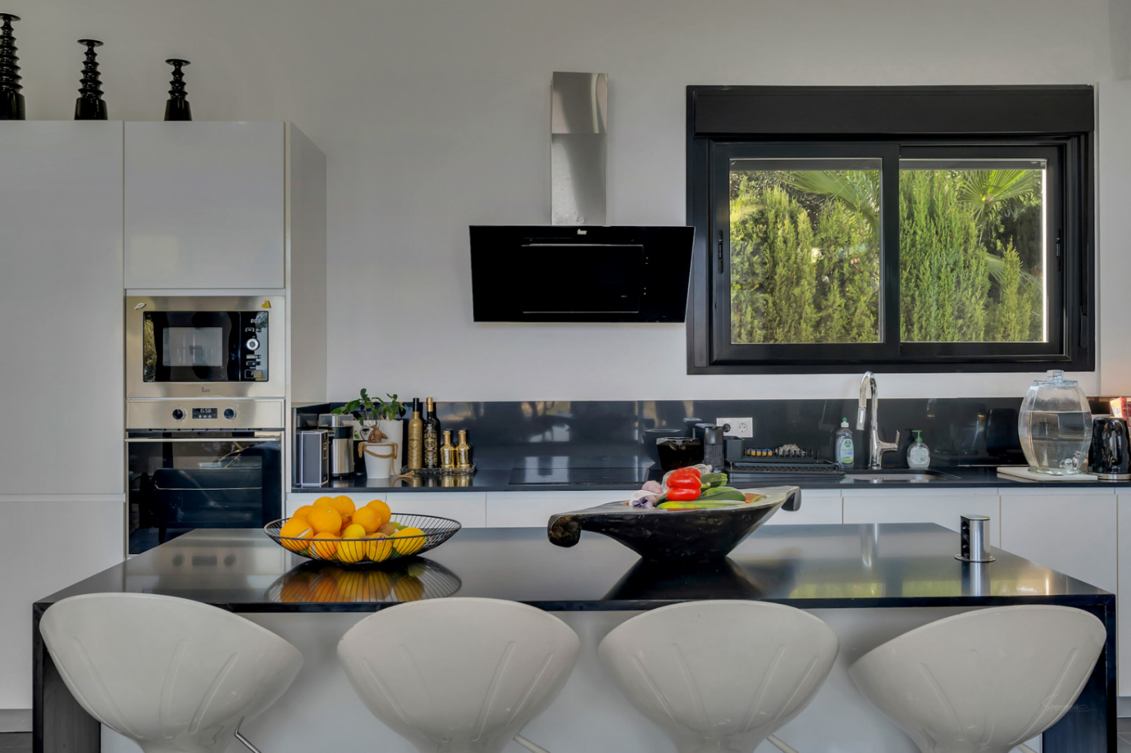 Cocina de concepto abierto con isla negra, taburetes de diseño blancos y ventanal con vistas directas al jardín mediterráneo.