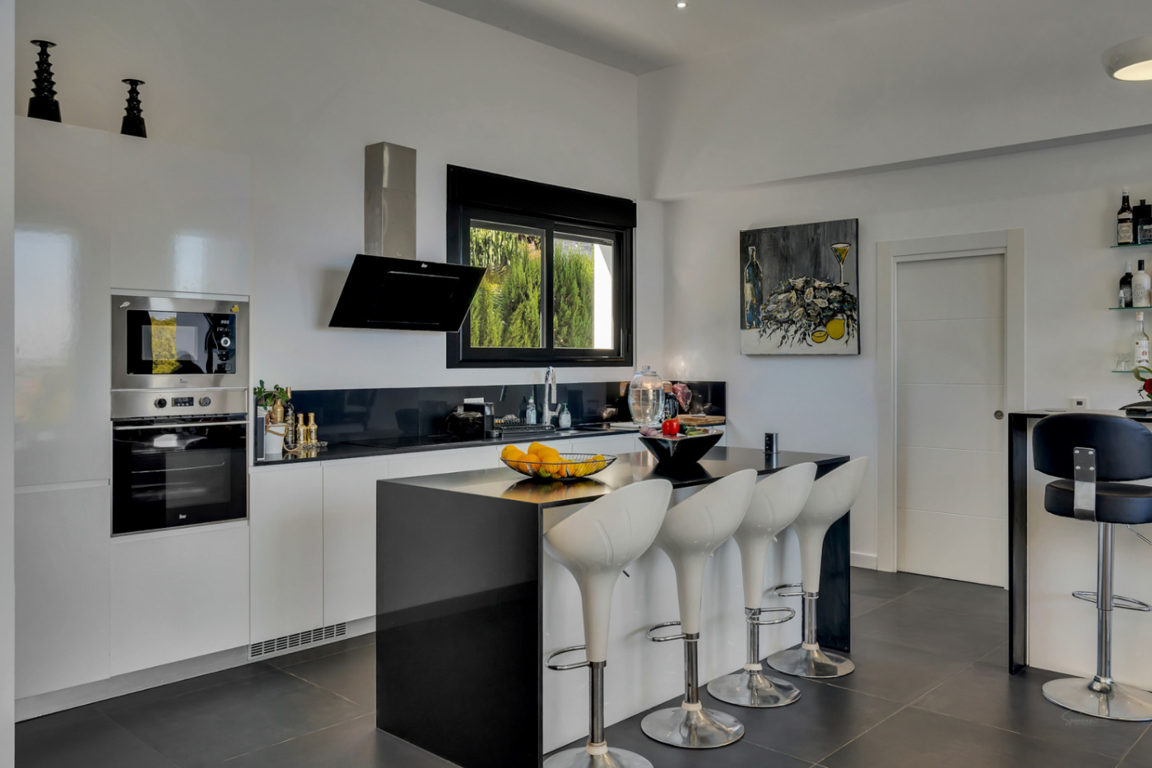Cocina de concepto abierto con isla negra, taburetes de diseño y electrodomésticos integrados. Destaca el contraste entre el mobiliario blanco y las encimeras oscuras.