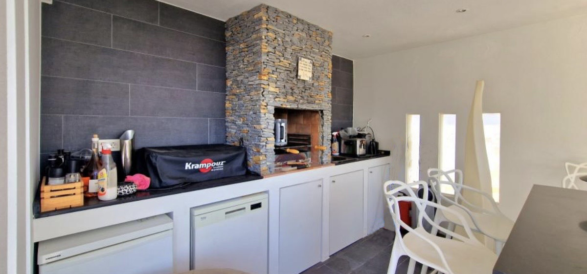 Cocina de verano equipada con barbacoa de obra revestida en piedra natural, encimera oscura y electrodomésticos integrados bajo muebles blancos.