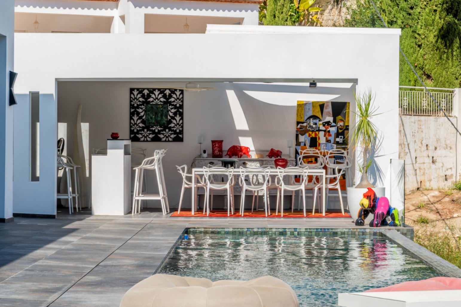 Cocina de verano y comedor exterior junto a la piscina, decorados con arte contemporáneo, mobiliario de diseño blanco y suelos de gres porcelánico.