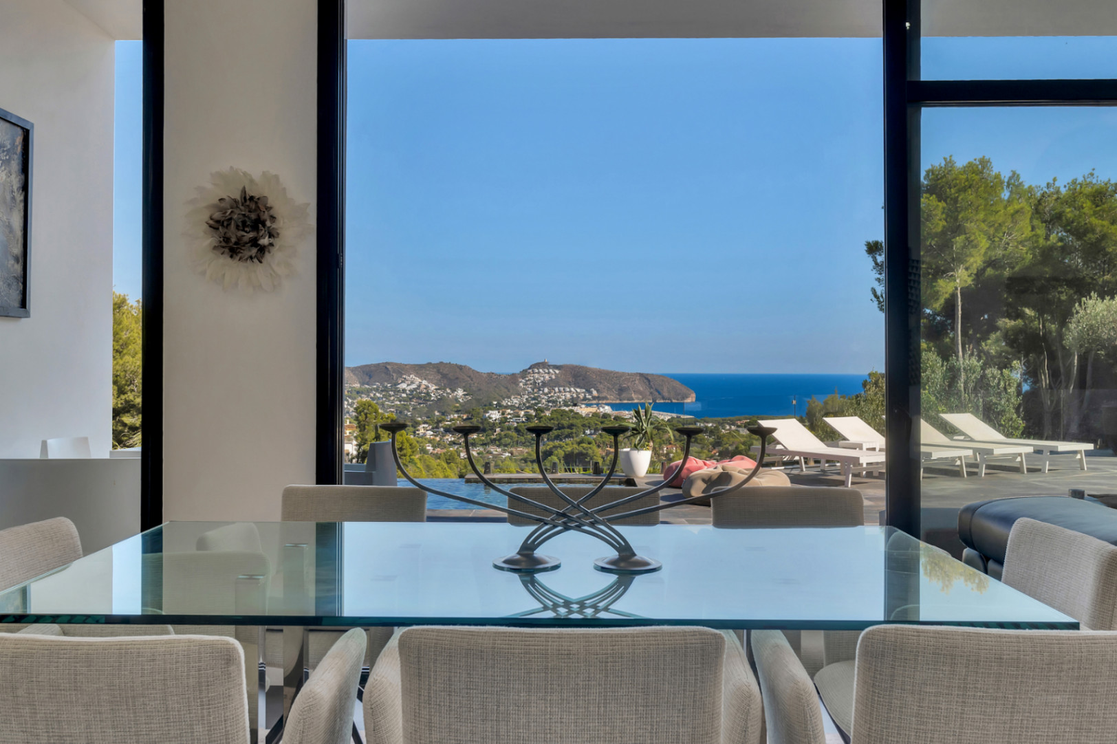 Comedor con mesa de cristal y sillas tapizadas, integrado visualmente con la terraza y las vistas panorámicas al mar Mediterráneo y la costa.