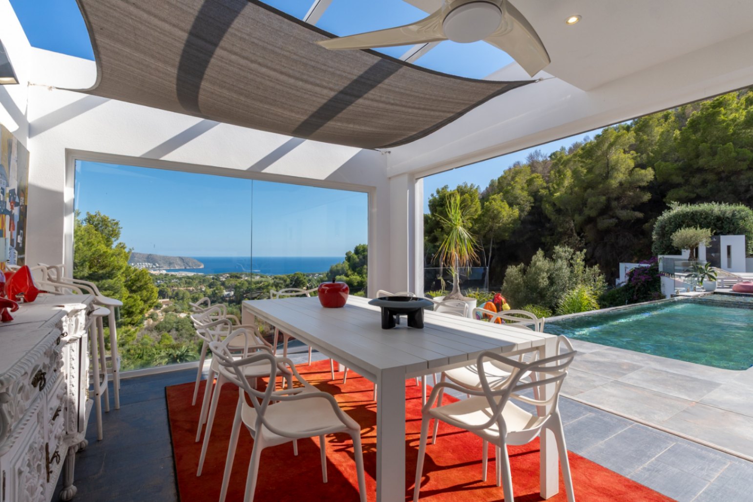 Comedor exterior acristalado con vistas al mar, equipado con mesa de diseño, alfombra roja y ventilador de techo junto a la piscina infinita.