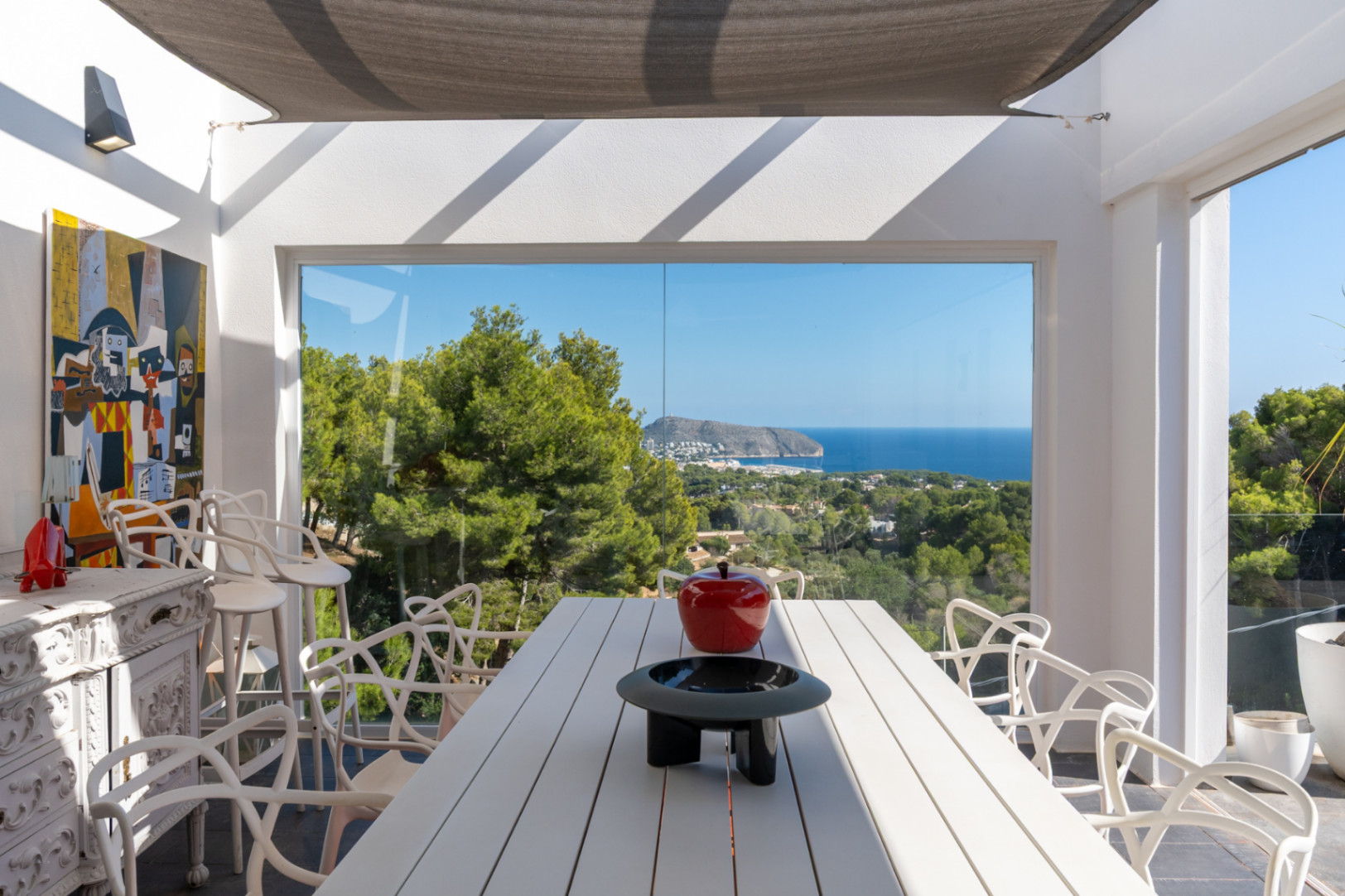Comedor exterior bajo toldo con mesa blanca y sillas de diseño, ofreciendo vistas panorámicas al mar y la costa de Moraira a través de grandes ventanales.