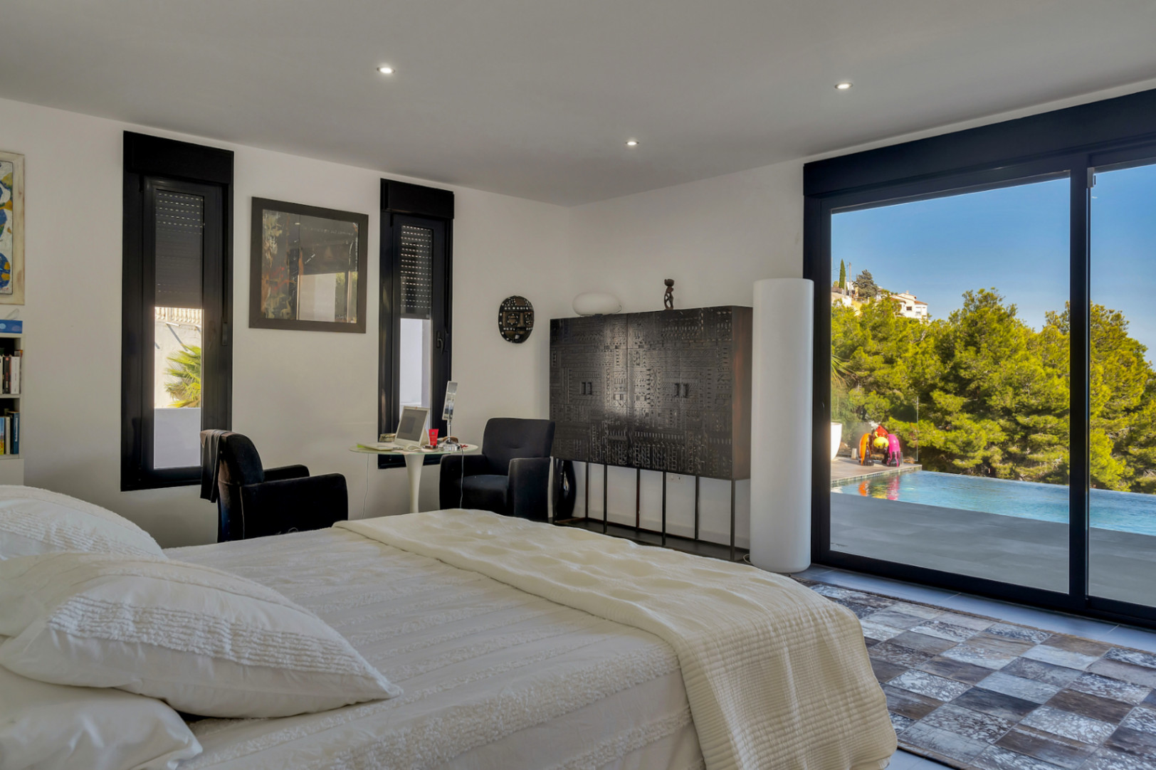 Dormitorio con acceso directo a la piscina y terraza. Destaca el mobiliario de diseño oscuro sobre paredes blancas, iluminación empotrada y vistas a la zona arbolada.