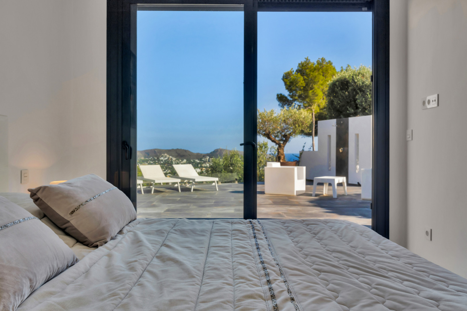 Dormitorio con acceso directo a la terraza mediante grandes ventanales. Vistas panorámicas a la montaña y zona exterior con mobiliario de diseño y suelos de piedra natural.