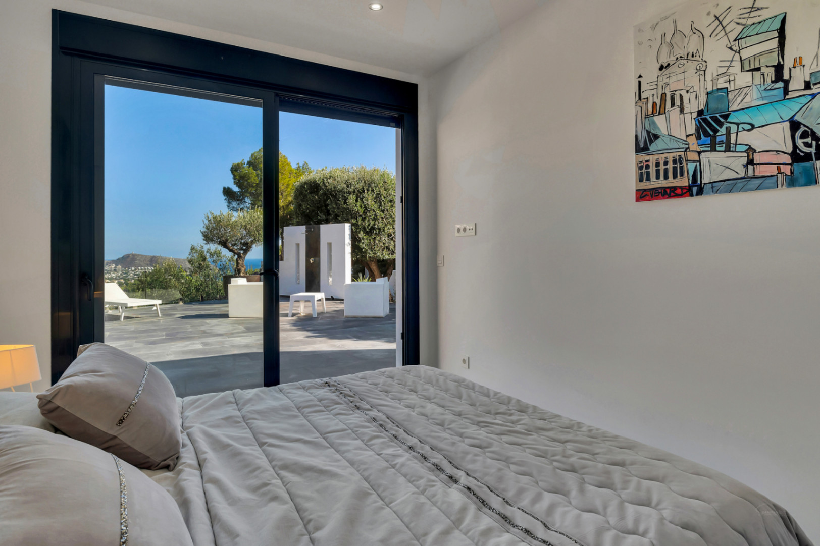 Dormitorio con acceso directo a la terraza mediante ventanales de carpintería negra, ofreciendo vistas al mar, mobiliario exterior blanco y suelos de piedra natural.