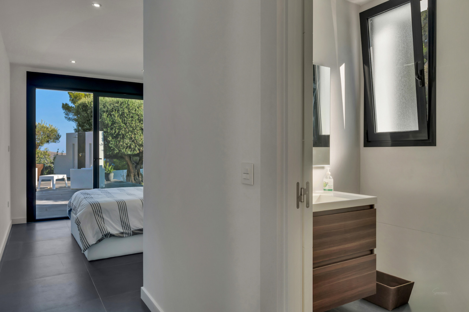 Dormitorio en suite con acceso directo a la terraza, suelos de gres oscuro y baño integrado con mueble de madera y ventana oscilobatiente.