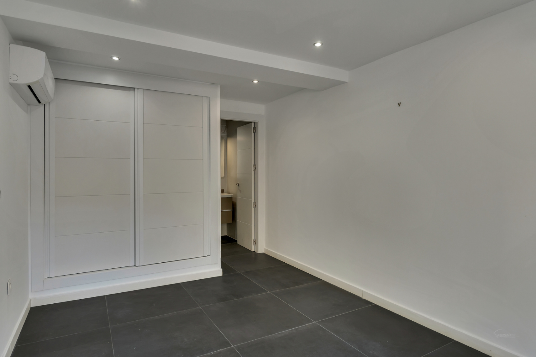 Dormitorio funcional con armario empotrado blanco, suelos de gres oscuro, aire acondicionado y acceso directo a baño en suite con iluminación empotrada.