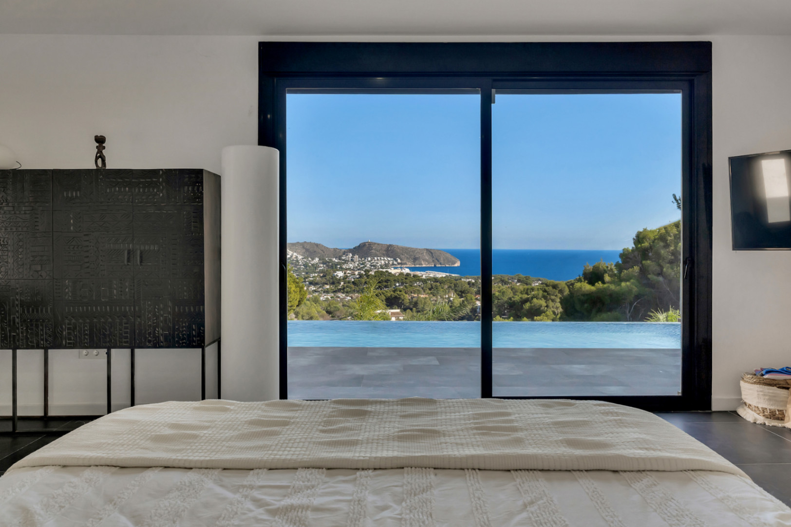 Dormitorio principal con vistas panorámicas al mar y la piscina infinity a través de ventanales. Incluye mobiliario de diseño y suelos de gres oscuro.