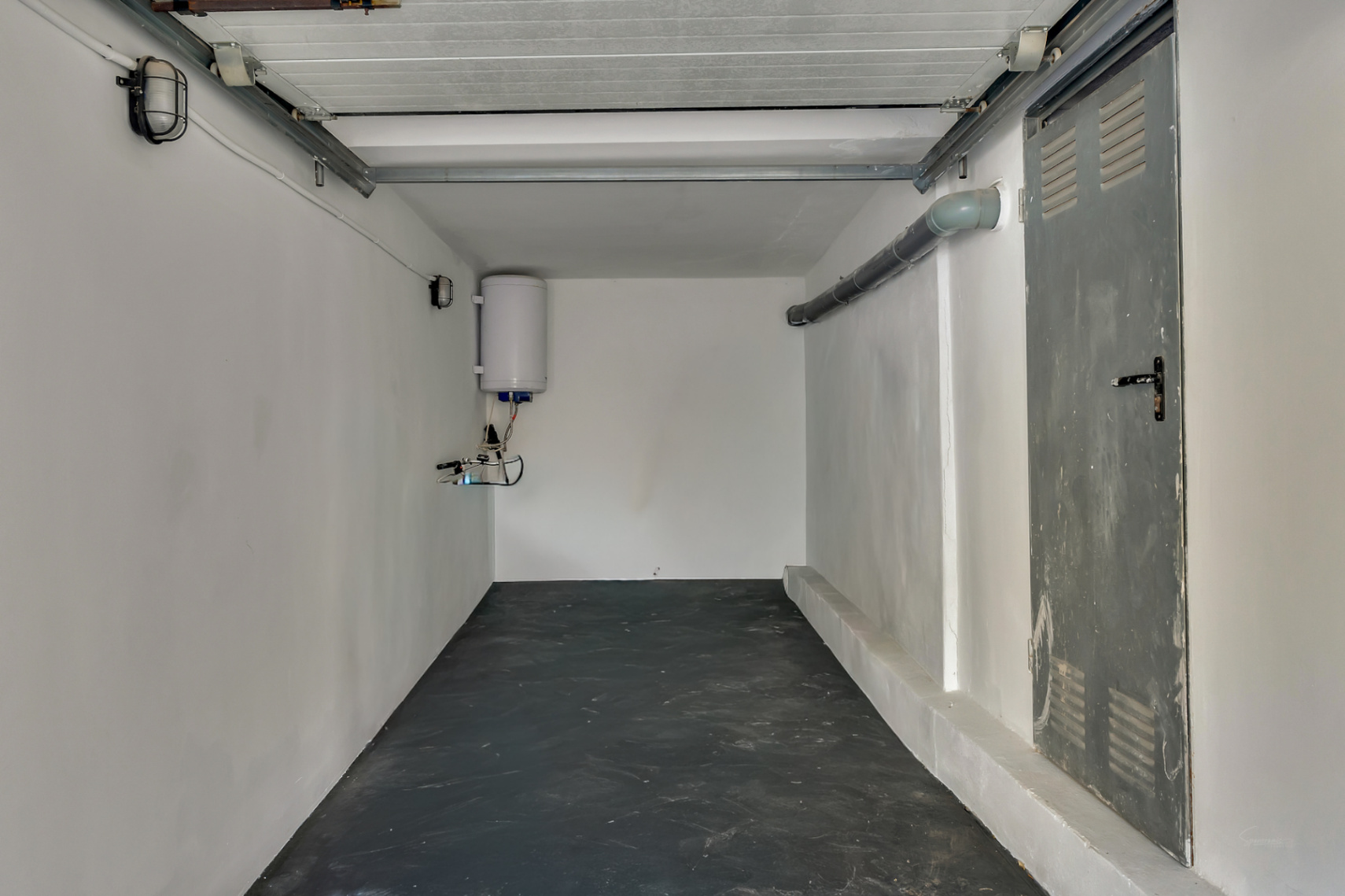 Garaje o trastero funcional con suelo oscuro, paredes blancas, termo eléctrico instalado y puerta metálica lateral con rejillas de ventilación.