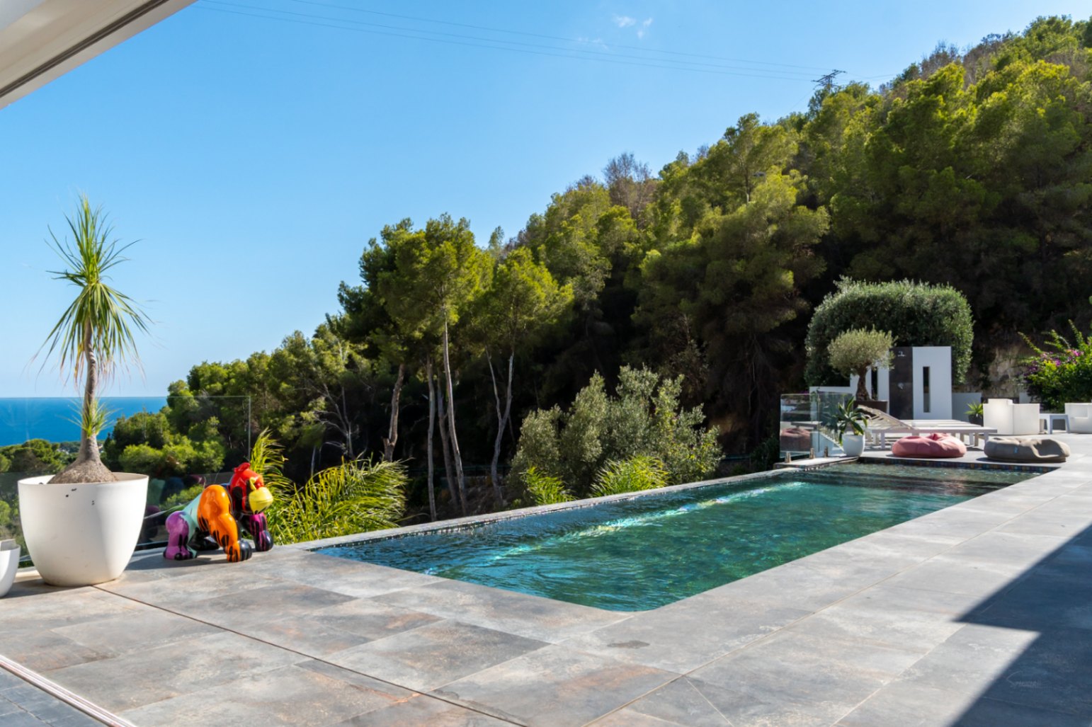 Piscina desbordante rodeada de terraza con baldosas de gran formato, vistas al mar y bosque mediterráneo, decorada con una escultura artística de un gorila.