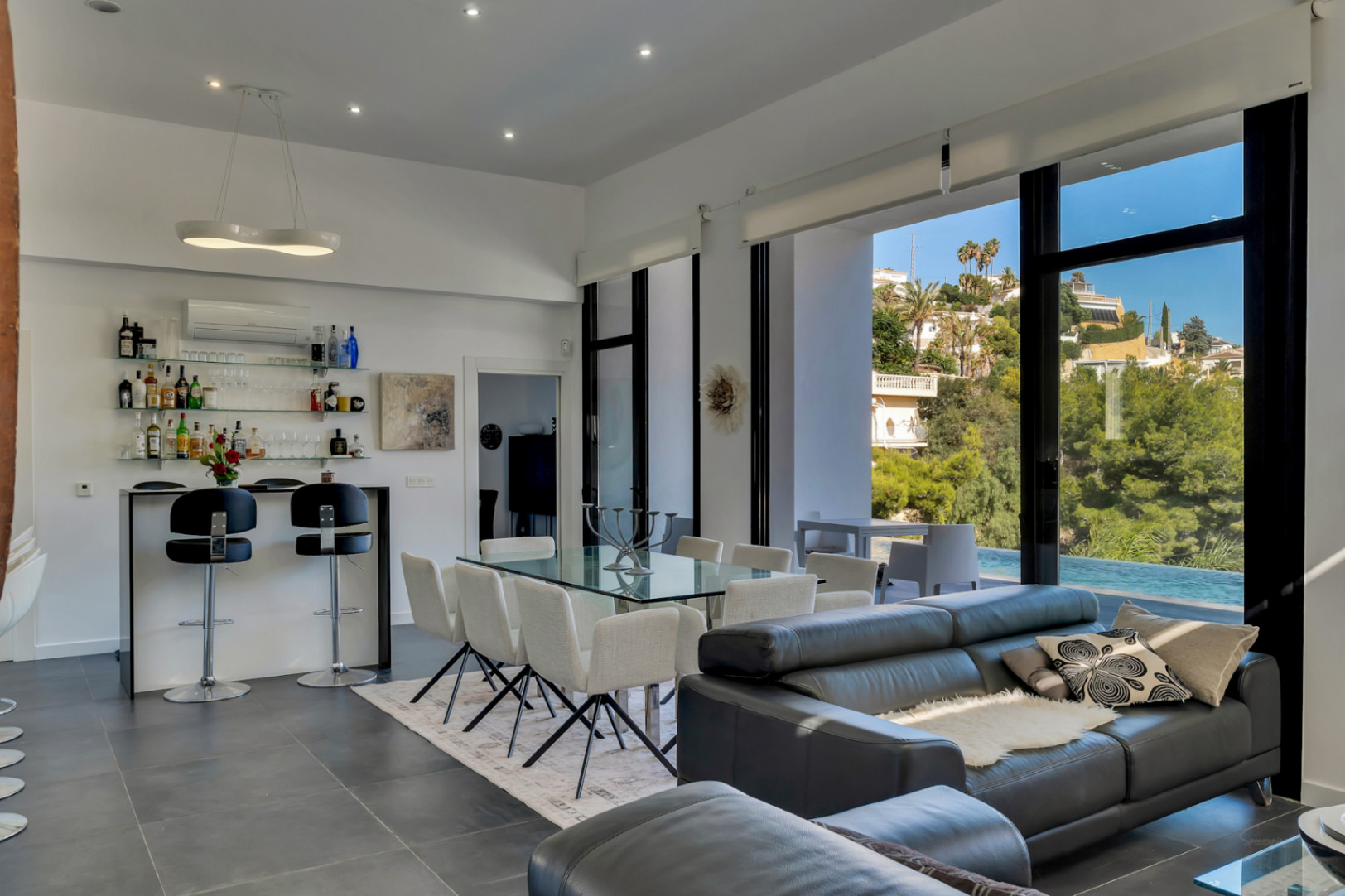 Salón-comedor diáfano con techos altos, zona de bar equipada y grandes ventanales que integran la terraza y la piscina con vistas al entorno natural.