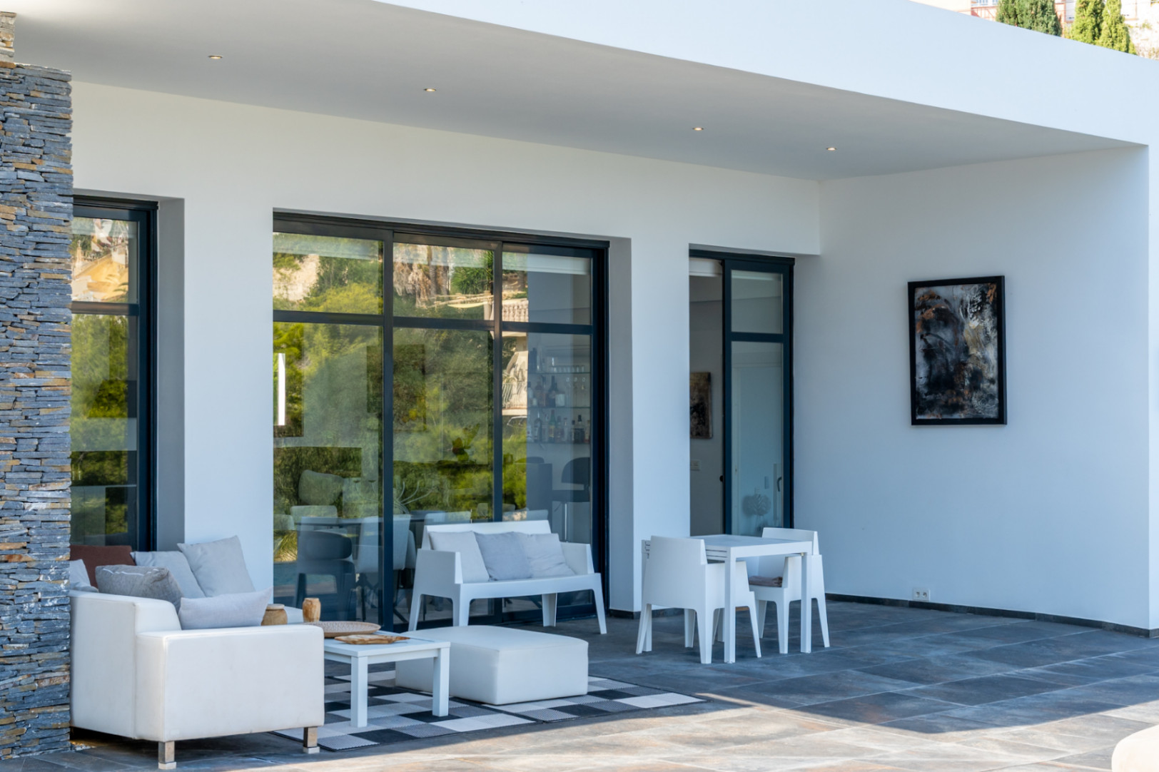 Terraza cubierta con suelos oscuros, mobiliario blanco de diseño y grandes ventanales de perfilería negra. Incluye un pilar de piedra natural y focos empotrados en el techo.