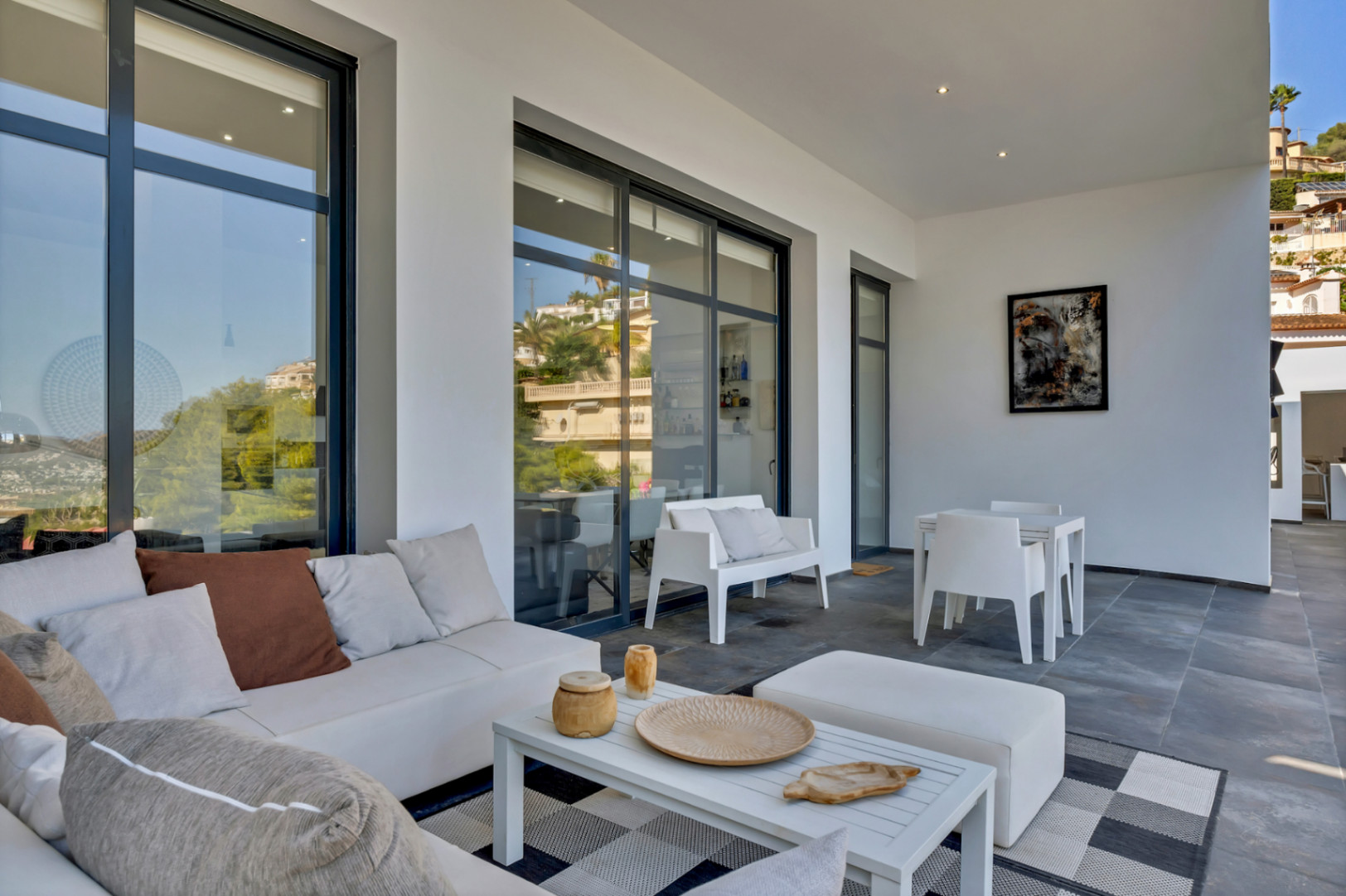 Terraza cubierta con zona de estar de diseño, mobiliario blanco, suelos de piedra gris y grandes ventanales que conectan con el interior de la villa.