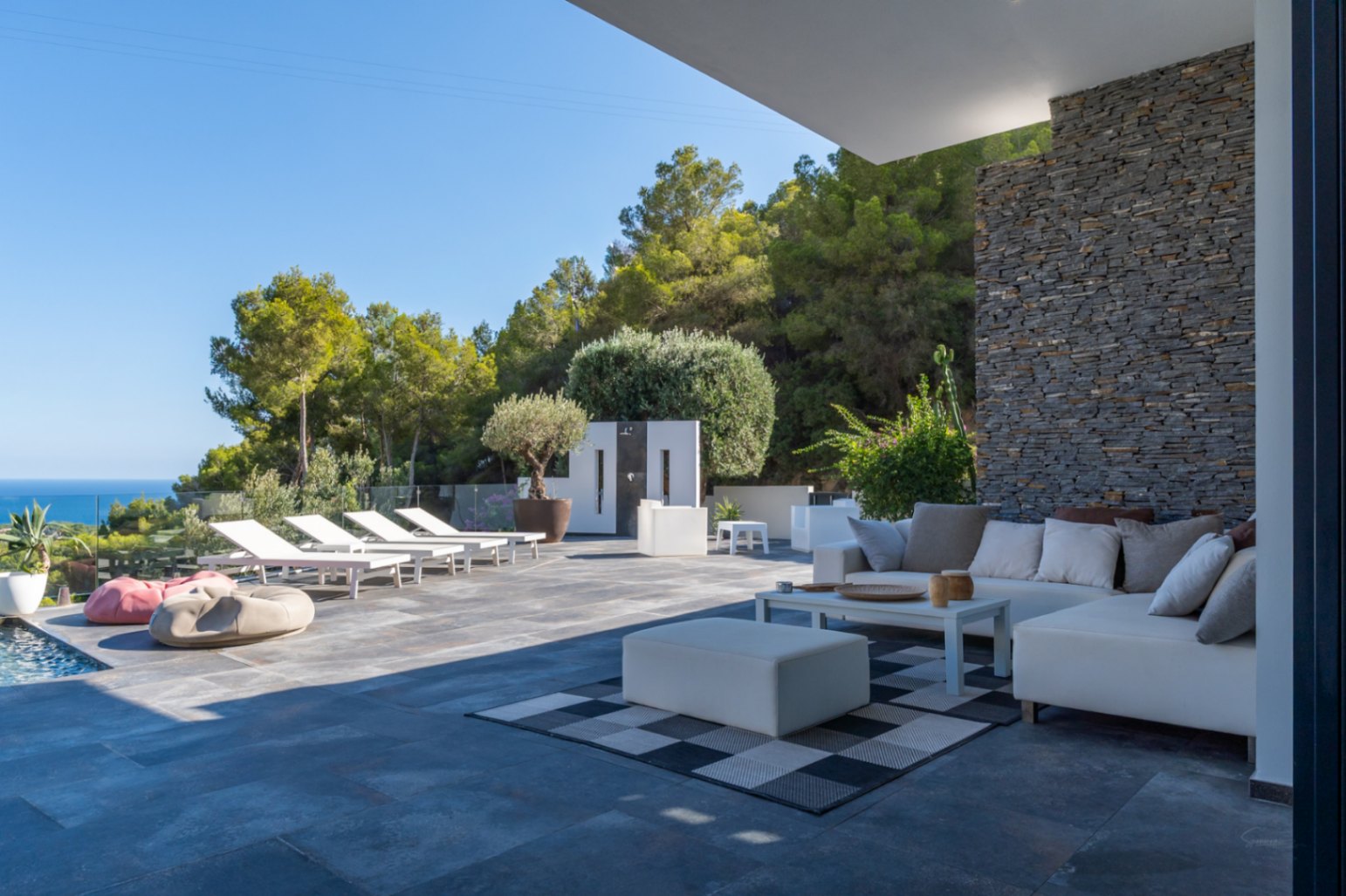 Terraza exterior con zona de estar, sofás modulares blancos y pared de piedra natural. Incluye solárium con tumbonas y vistas despejadas al mar y la montaña.
