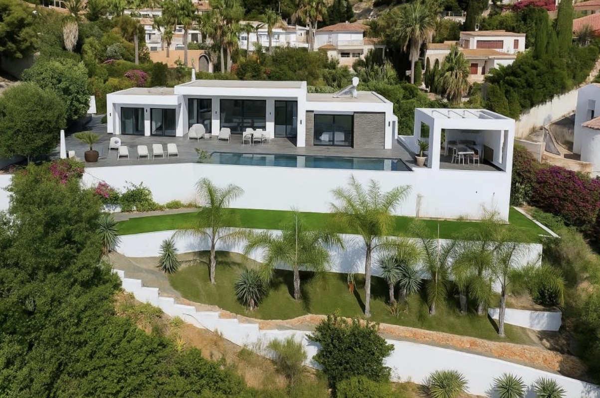 Vista exterior de la villa moderna con fachada blanca, piscina longitudinal y amplias terrazas. El jardín escalonado integra palmeras y zonas de césped frente a la arquitectura lineal.