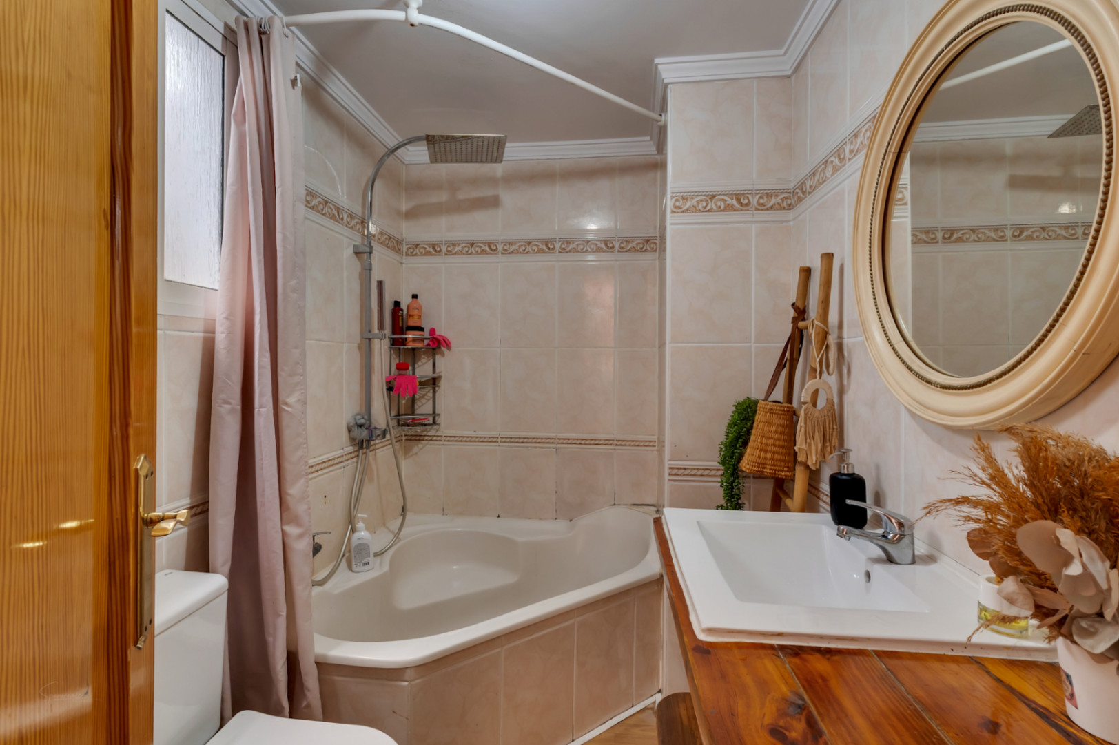 Baño completo con bañera de rincón, lavabo sobre encimera de madera rústica y espejo circular decorativo. Paredes alicatadas con cenefa clásica y detalles en madera.