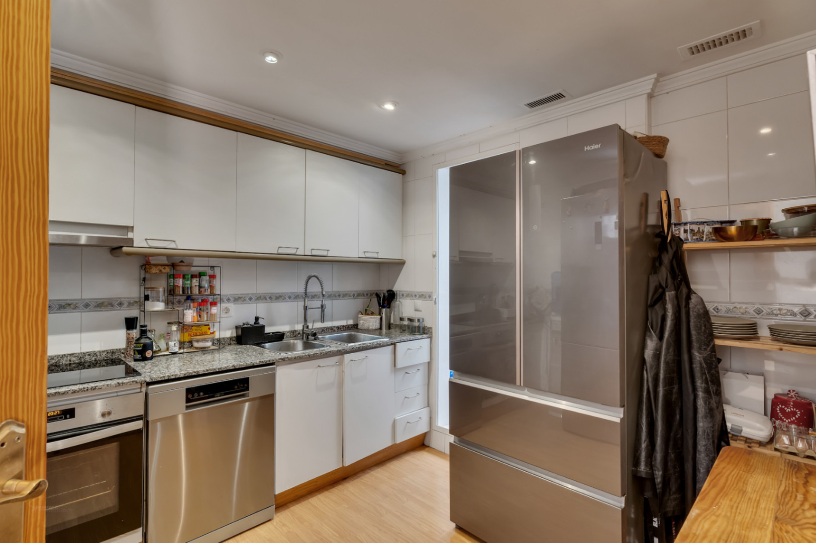 Cocina funcional con armarios blancos, encimera de granito y electrodomésticos de acero inoxidable, incluyendo un frigorífico de gran capacidad y lavavajillas integrado.
