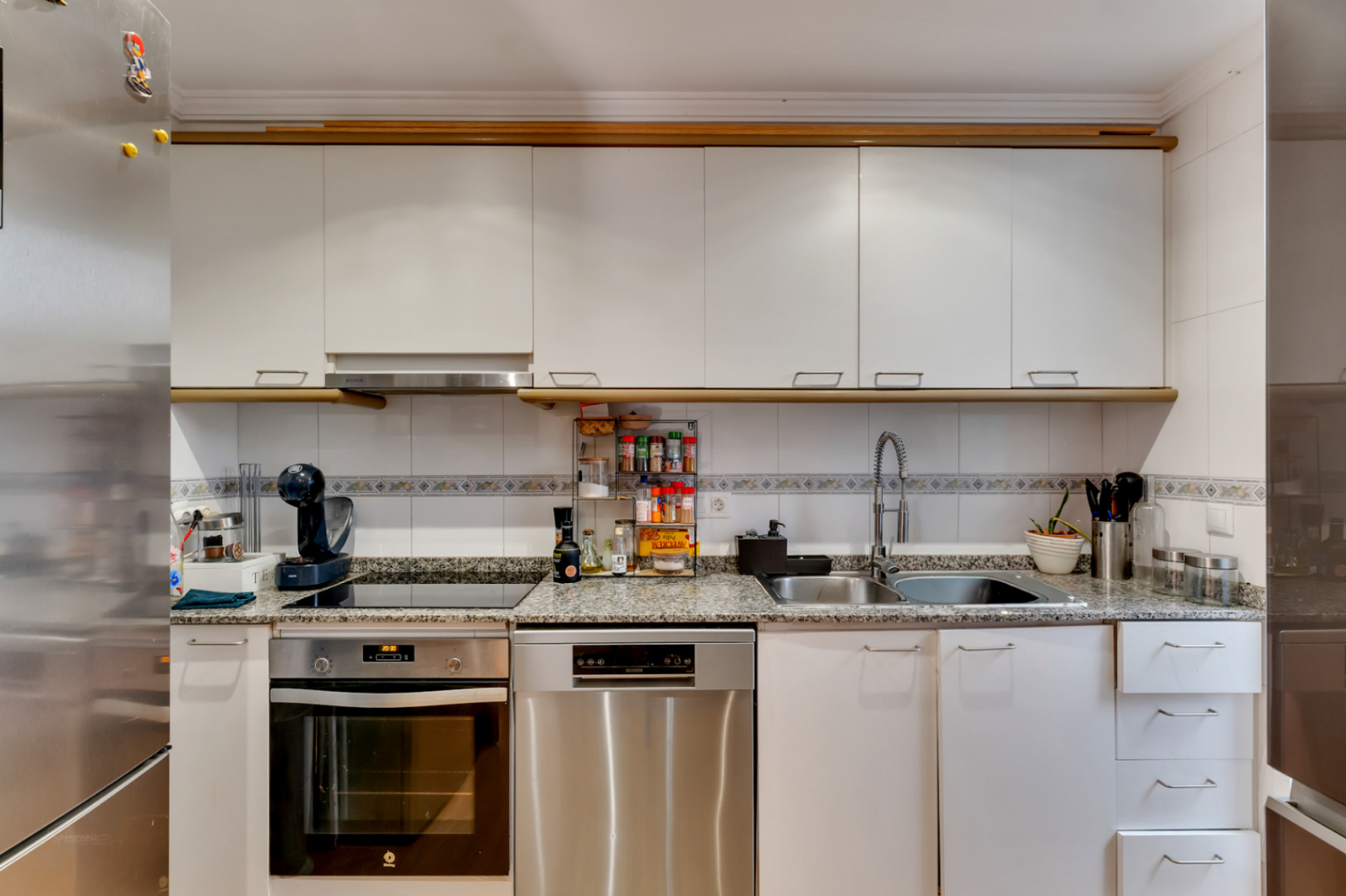 Cocina funcional con encimera de granito, armarios blancos y electrodomésticos de acero inoxidable. Incluye placa vitrocerámica, horno y lavavajillas integrados.