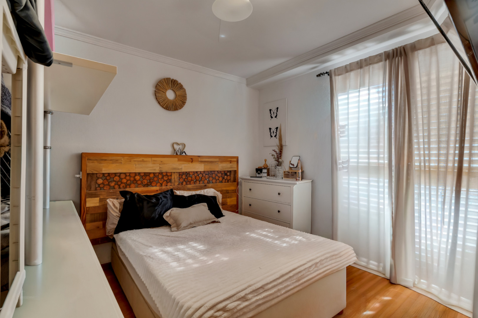 Dormitorio acogedor con cama doble, cabecero artesanal de madera y suelos laminados. Dispone de cómoda blanca y gran ventanal con cortinas que filtran la luz natural.