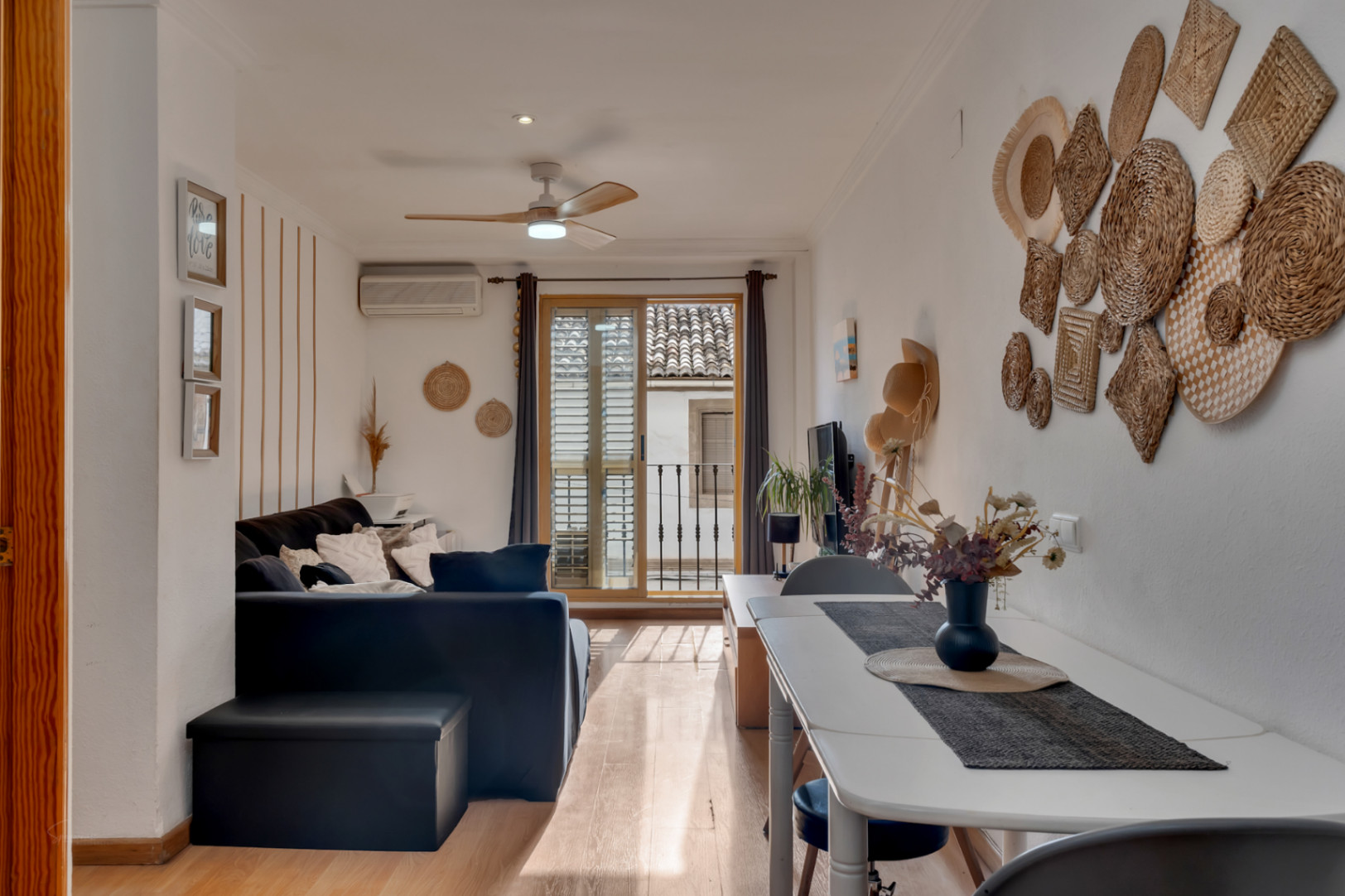 Salón comedor luminoso con suelos de madera, ventilador de techo y acceso a un balcón con vistas al casco antiguo. Decoración de estilo mediterráneo con detalles artesanales.