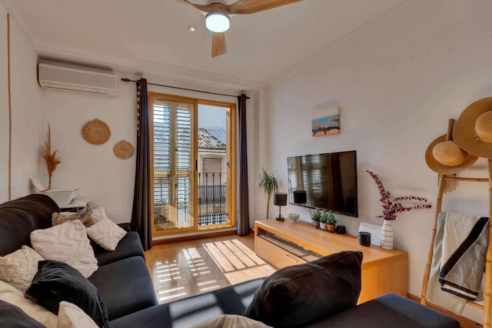 Salón luminoso con suelo de madera, aire acondicionado y salida a balcón con contraventanas. Incluye ventilador de techo y zona de TV con mobiliario funcional.