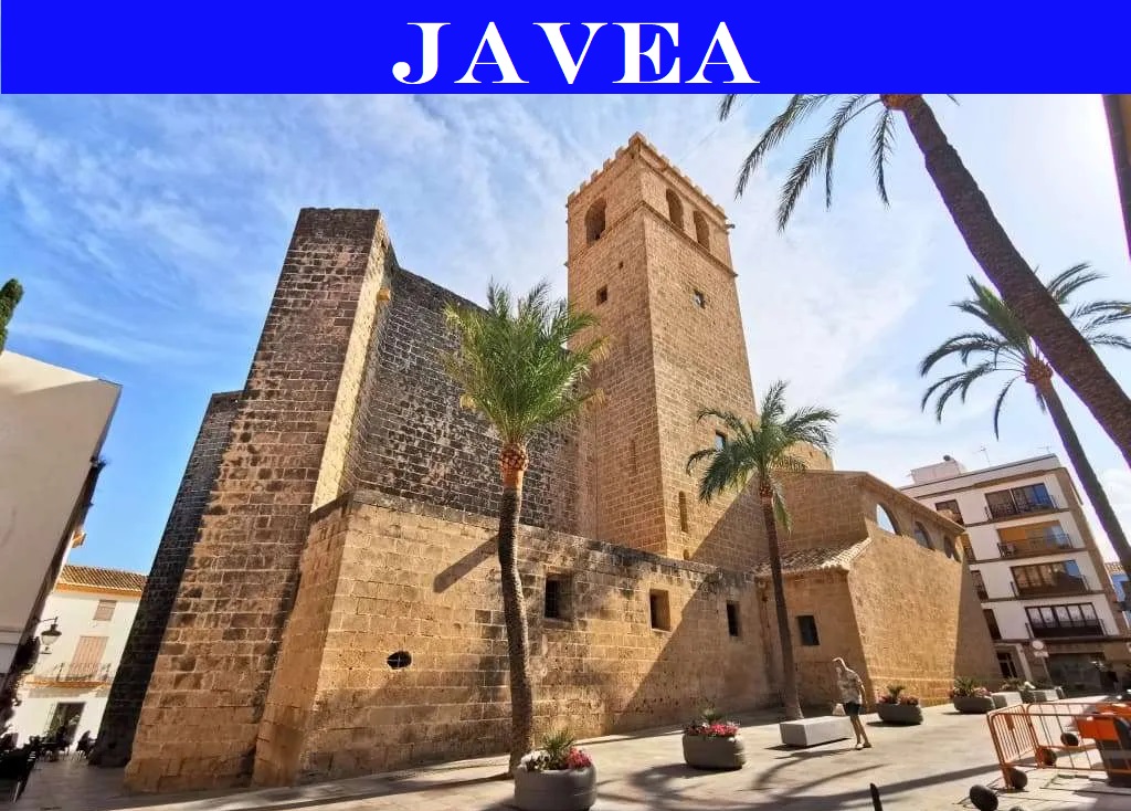 Vista exterior de la Iglesia de San Bartolomé en el casco antiguo de Jávea, destacando su arquitectura de piedra tosca y el entorno de plazas peatonales con palmeras.
