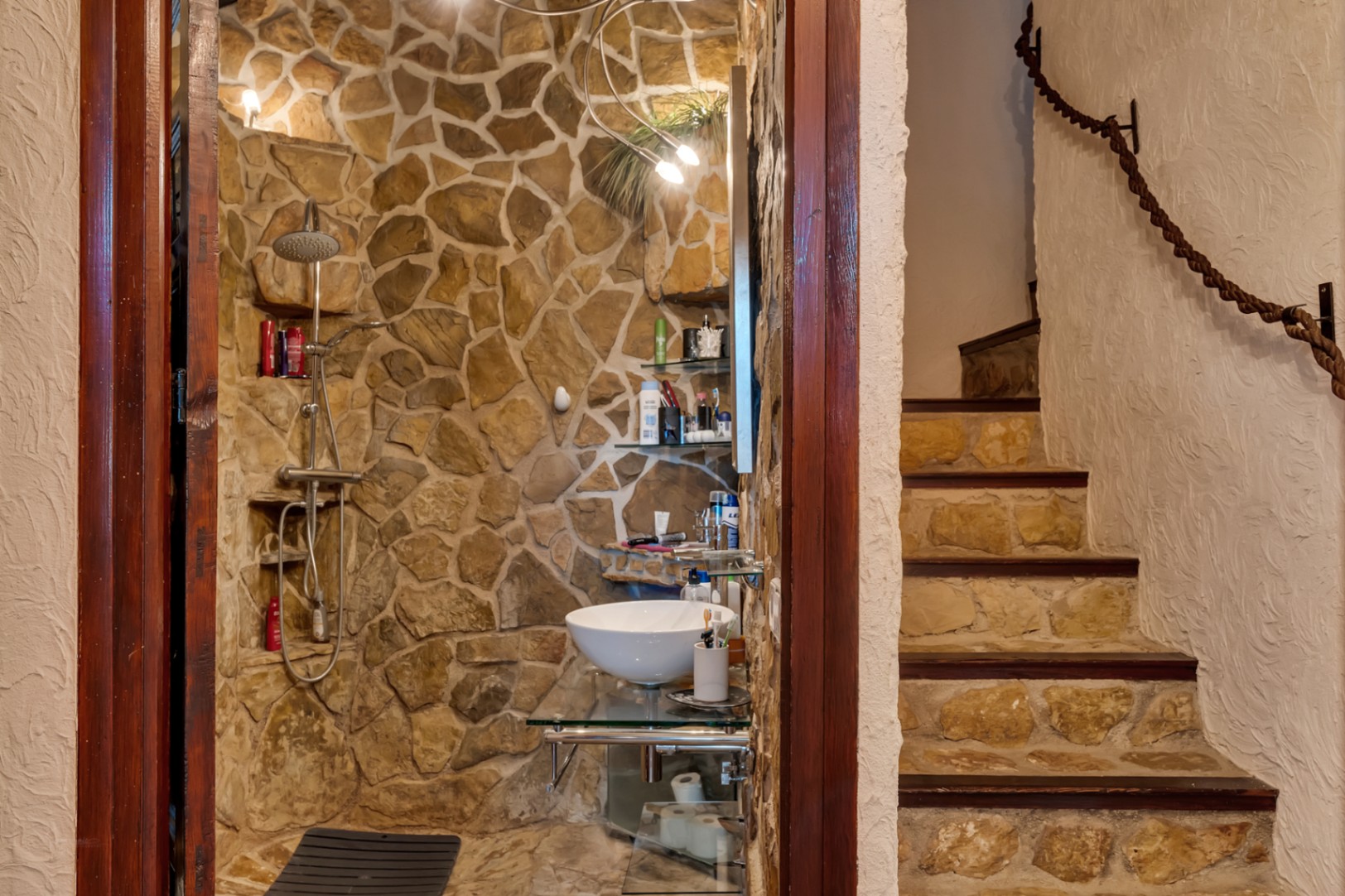 Baño de estilo rústico con paredes de piedra natural, ducha integrada y lavabo sobre cristal, junto a una escalera con pasamanos de cuerda.