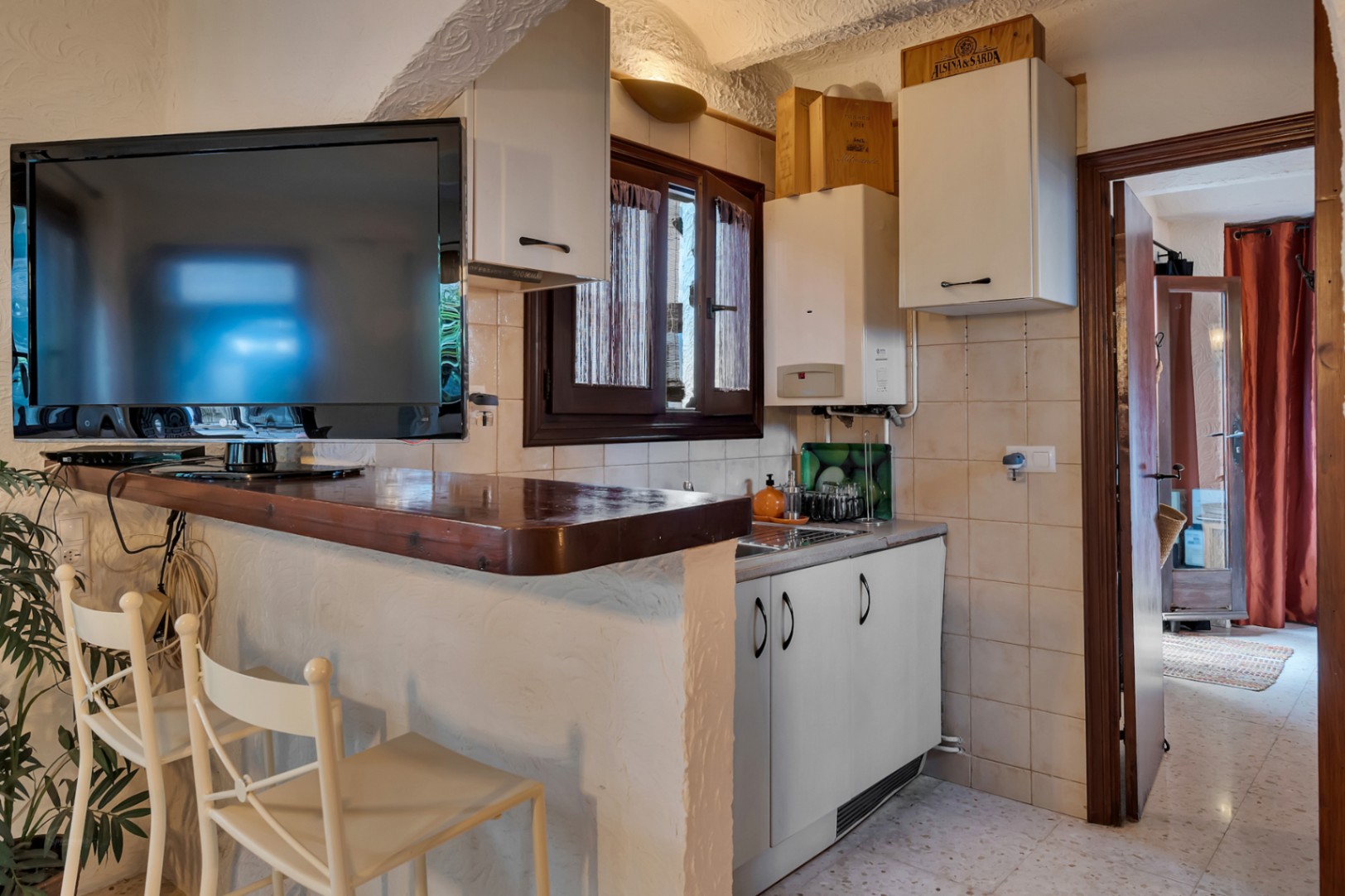 Cocina compacta con barra de madera, armarios blancos y televisor integrado. El espacio presenta paredes de yeso texturizado y acceso directo a las estancias contiguas.