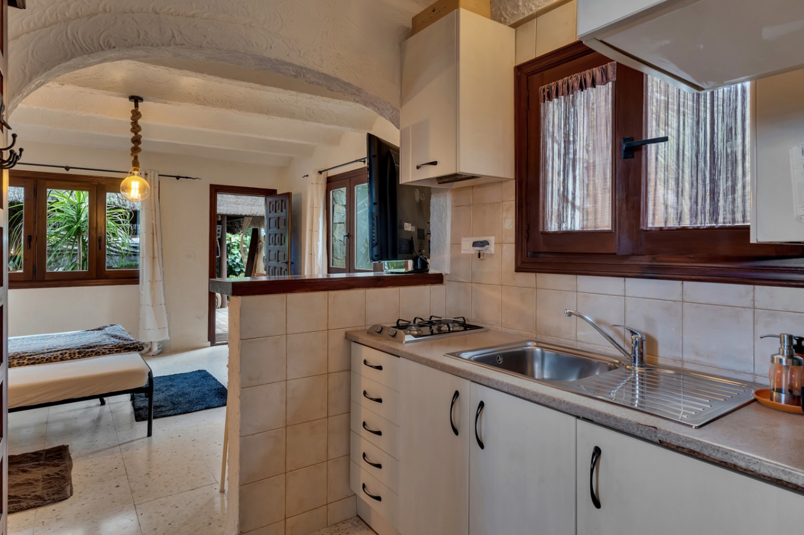 Cocina compacta funcional con encimera de piedra, fogones de gas y fregadero de acero inoxidable, integrada bajo un techo abovedado de estilo mediterráneo.