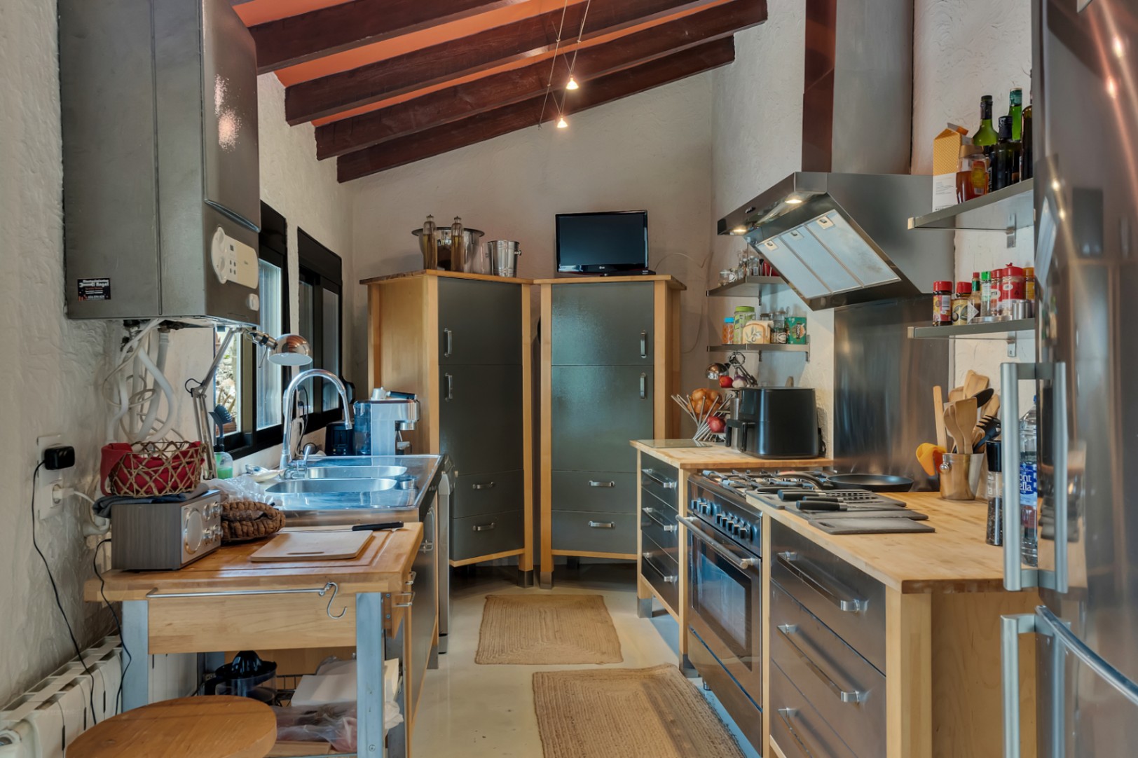 Cocina de estilo industrial con vigas de madera vistas, encimeras de madera maciza y electrodomésticos de acero inoxidable bajo un techo inclinado tradicional.