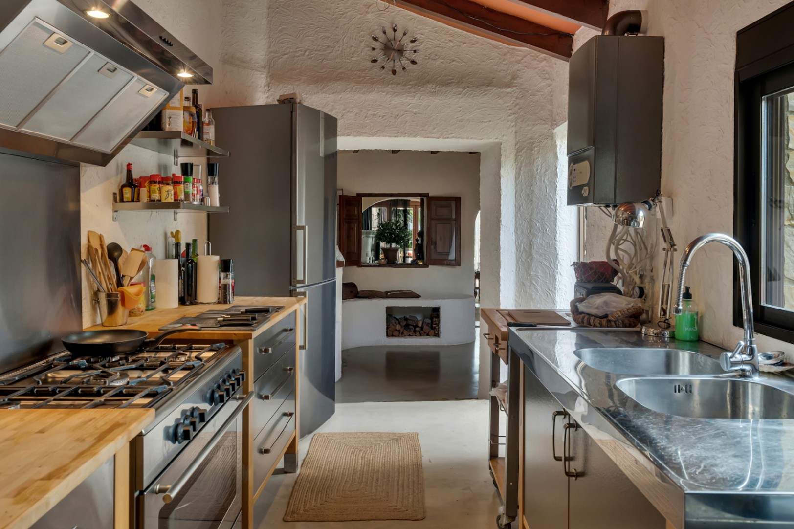 Cocina rústica moderna con encimeras de madera y acero inoxidable, paredes de textura blanca y vistas a una estancia con chimenea de leña.