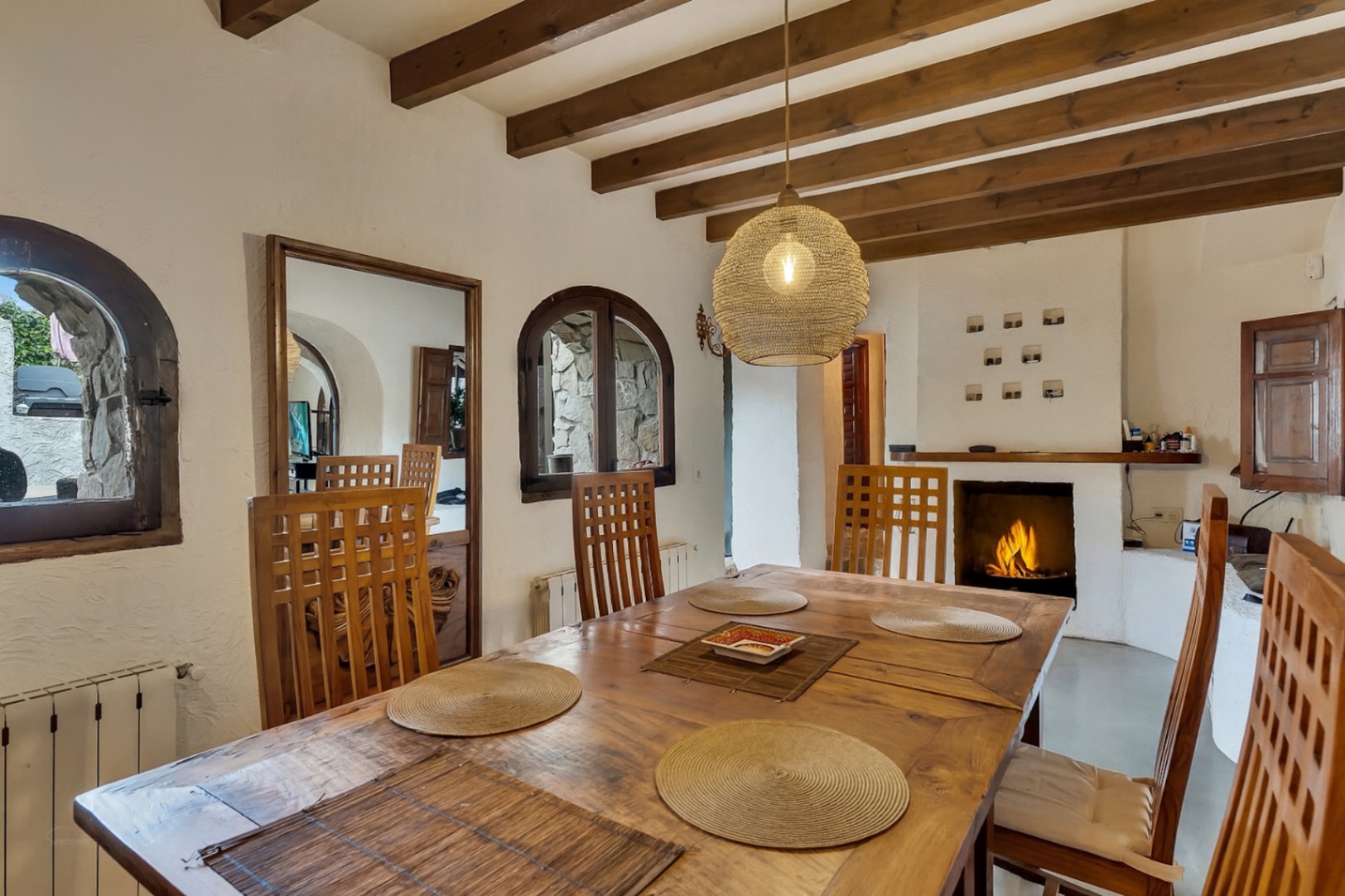 Comedor rústico con vigas de madera vista, mesa de madera maciza y chimenea de obra encendida. Destacan las ventanas de arco y la lámpara de fibras naturales.