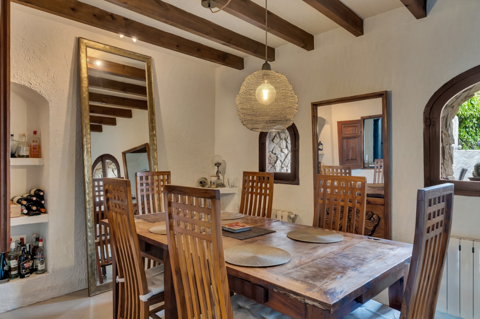Comedor rústico con vigas de madera vista, mesa de madera maciza y lámpara colgante de fibra natural. Incluye un gran espejo de pie y hornacinas integradas para almacenaje.