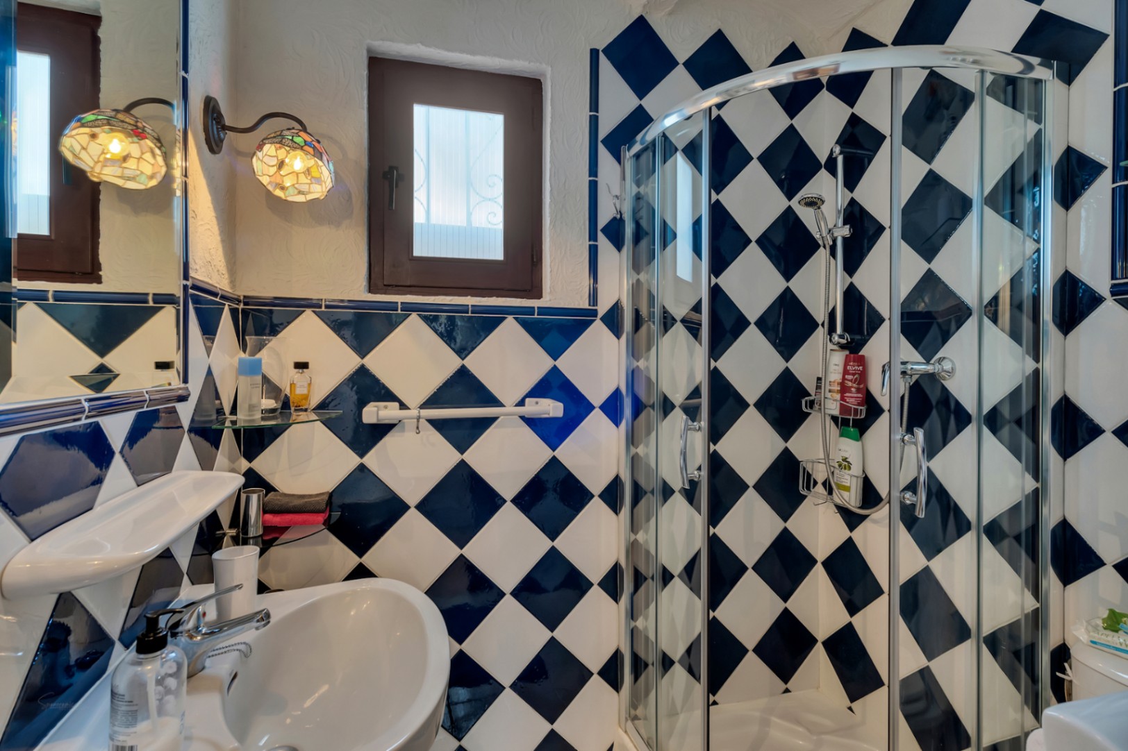 Cuarto de baño con azulejos en damero azul y blanco, ducha de esquina con mampara curva de cristal y apliques decorativos de estilo Tiffany.