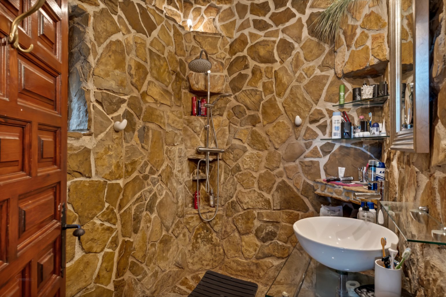 Cuarto de baño de estilo rústico con paredes de piedra natural, ducha integrada con grifería cromada y lavabo sobre encimera de cristal.