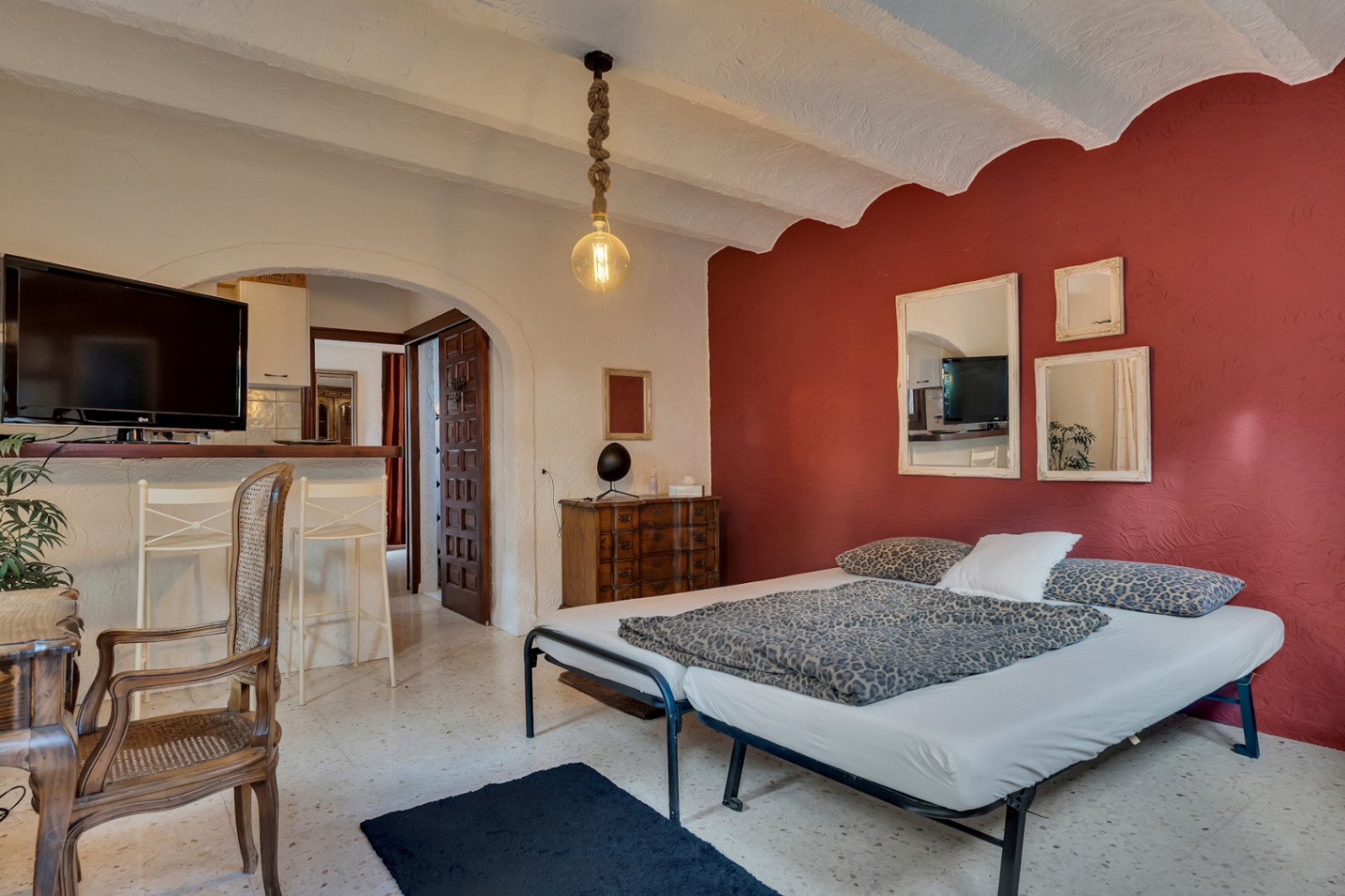 Dormitorio con pared de acento roja, techos abovedados blancos y suelos de terrazo. Incluye zona de estudio con escritorio de madera y una barra abierta hacia la cocina.