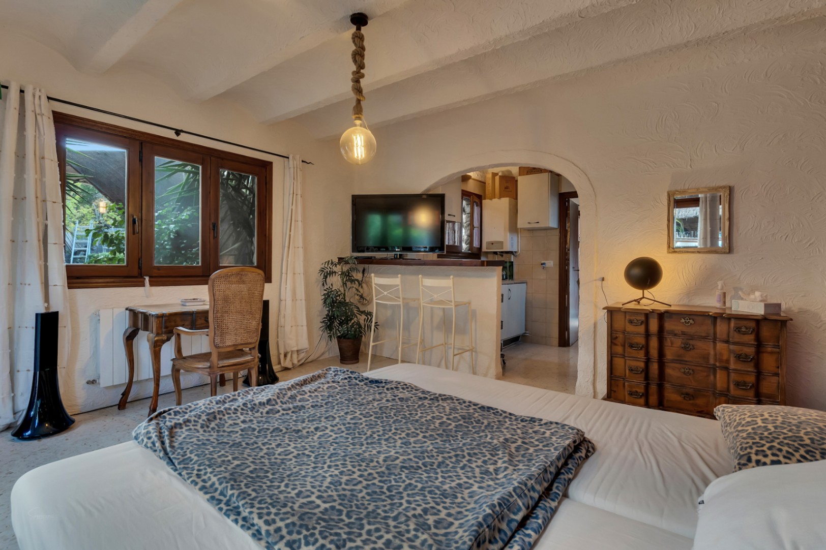 Dormitorio con zona de trabajo y acceso mediante arco a una cocina integrada con barra. Destacan las vigas vistas y las paredes de textura rústica mediterránea.