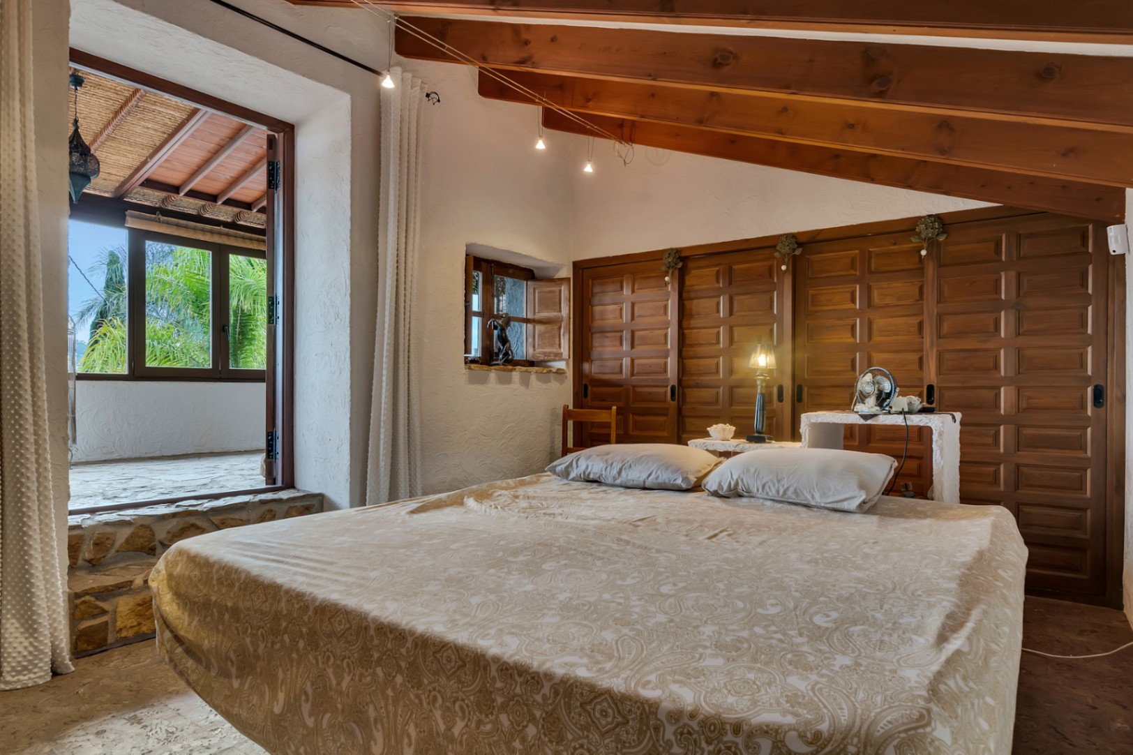 Dormitorio rústico con techos de vigas de madera inclinadas, armarios empotrados de madera tallada y acceso directo a una terraza cubierta con vistas al jardín.