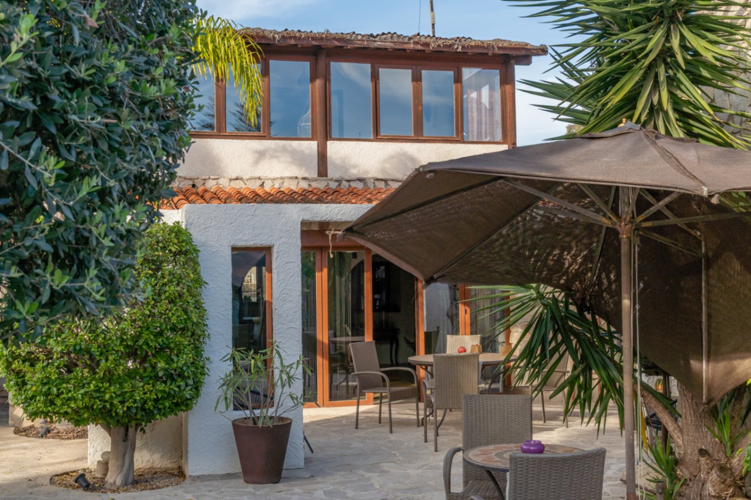 Fachada de la villa con terraza acristalada en la planta superior y zona de estar exterior pavimentada en piedra, rodeada de vegetación mediterránea.