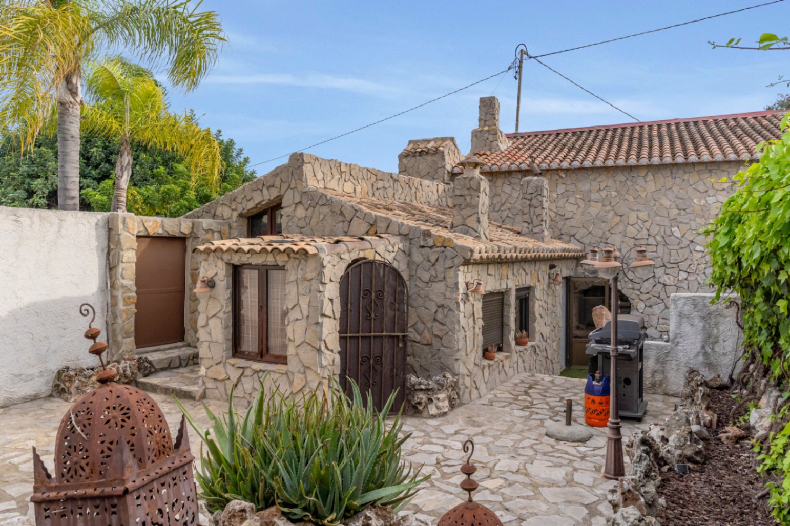 Fachada de piedra natural con arcos de hierro forjado y patio pavimentado. Incluye zona de barbacoa y detalles decorativos en un entorno mediterráneo tradicional.