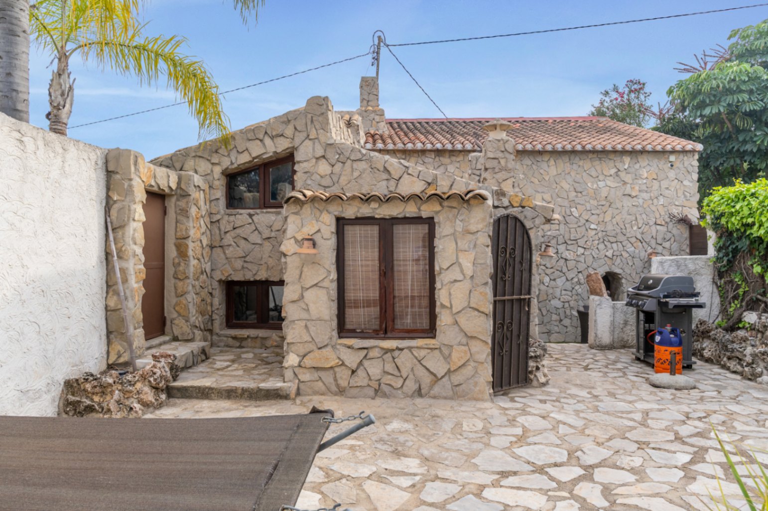 Fachada de piedra natural con carpintería de madera oscura y patio pavimentado. Incluye zona de barbacoa exterior y detalles arquitectónicos rústicos tradicionales.