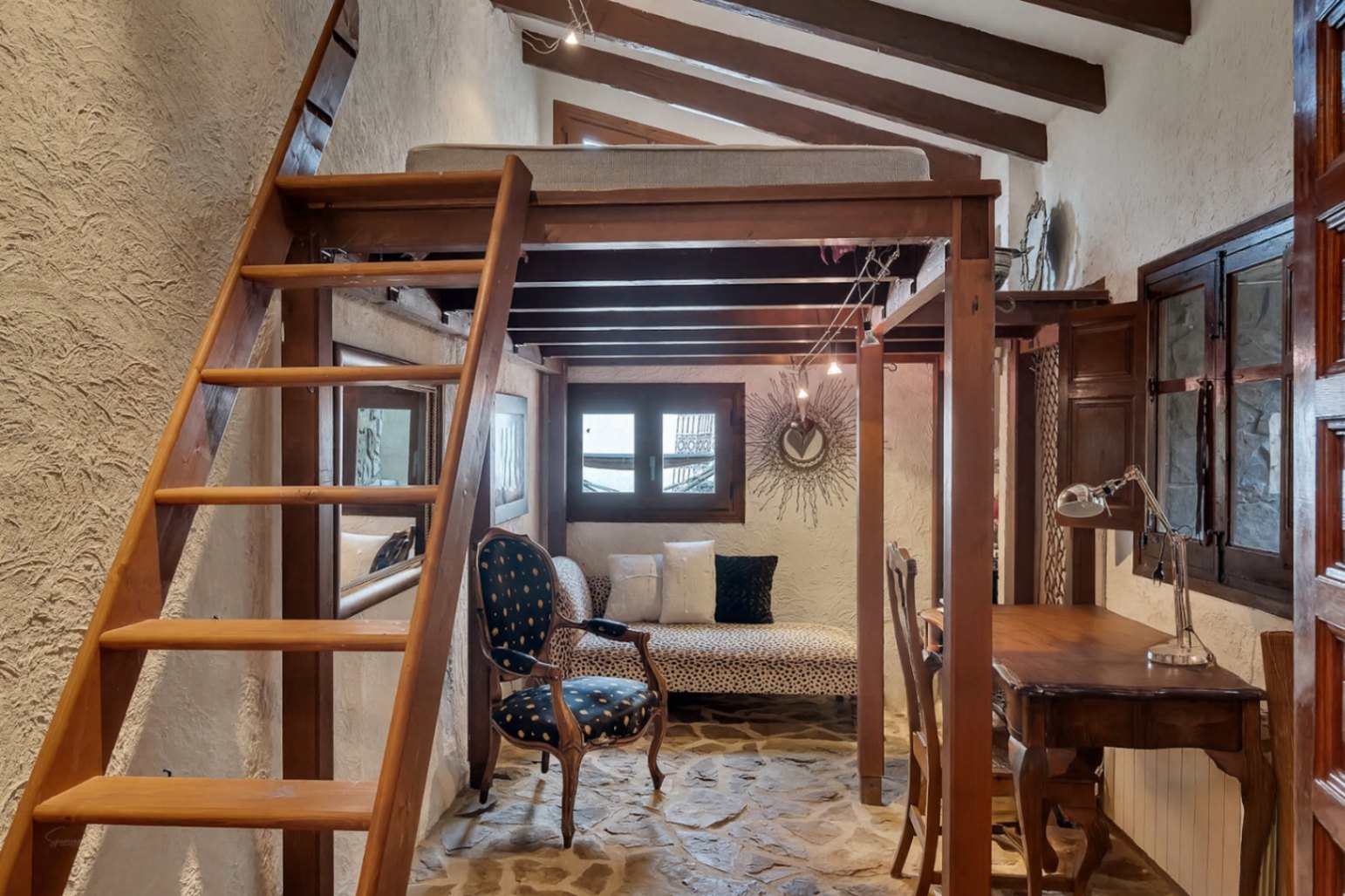 Habitación de estilo rústico con altillo de madera, suelos de piedra natural y vigas vistas. Incluye zona de estar inferior y escritorio bajo una estructura funcional.