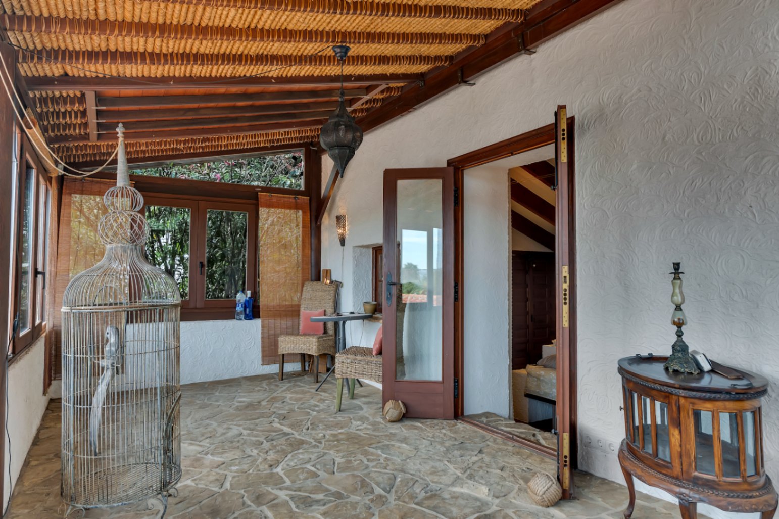 Naya acristalada con suelo de piedra natural, techo de vigas de madera con cañizo y mobiliario rústico que conecta el jardín con el interior de la vivienda.