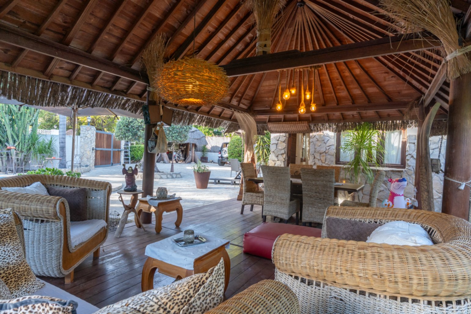 Pérgola de estilo balinés con estructura de madera vista, zona de estar con sofás de mimbre y comedor exterior sobre suelo de tarima oscura.