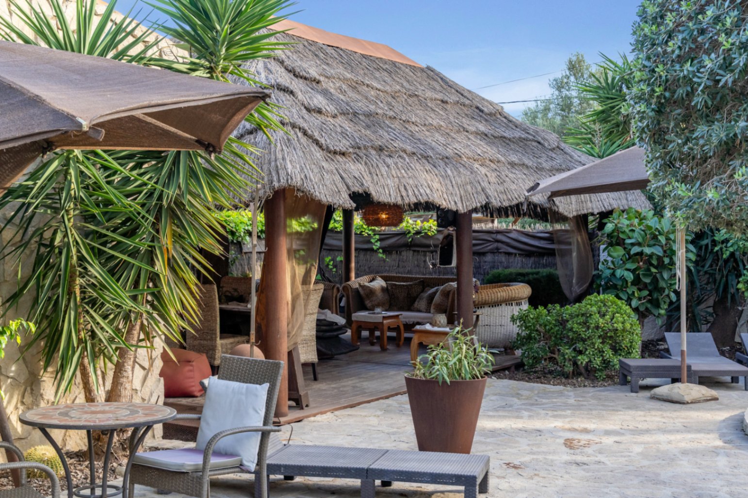 Pérgola de estilo balinés con techo de brezo y tarima de madera, rodeada de una terraza de piedra natural y vegetación mediterránea autóctona.