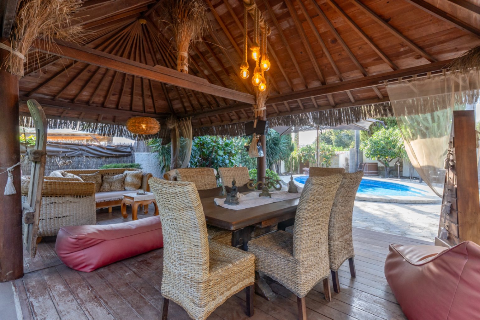 Pérgola de madera de estilo balinés con suelos de tarima, mobiliario de mimbre y vistas directas a la piscina privada.