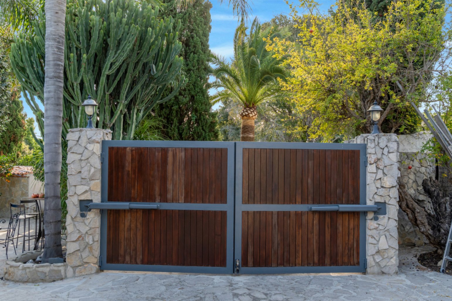 Puerta de acceso automática con paneles de madera y marco metálico, flanqueada por pilares de piedra natural y rodeada de vegetación mediterránea madura.