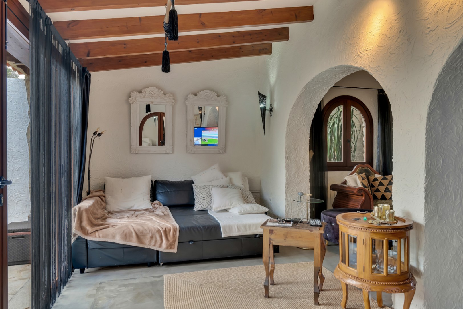 Salón acogedor con vigas de madera vistas, paredes de textura rústica y arcos tradicionales que conectan los espacios. Incluye mobiliario de cuero y detalles en madera natural.