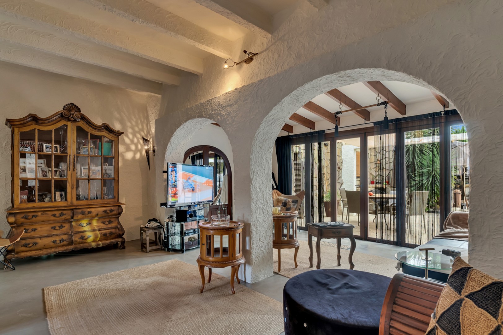 Salón de estilo mediterráneo con arcos de escayola, techos de vigas vistas y una vitrina clásica de madera. Grandes ventanales conectan el interior con la zona exterior.